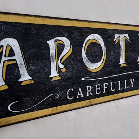 Close-up of the off-white lettering and gold accents on the rustic black background of the Apothecary sign.